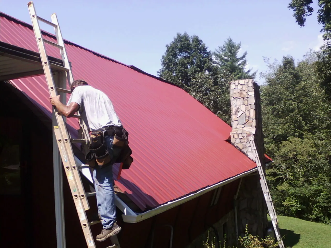 Roofer on a ladder beside a red metal roof and stone chimney — Soffit & Fascia Repair by Roofing Brothers in Fall City